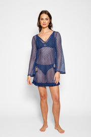 Brunette model facing front, wearing Navy Zuma Tunic with a sheer chevron crochet design, V-neckline with tassel trim, fringe hem, bell sleeves, and gold tassel accents. Koy Resort affordable vacation, cruise, and resort-wear.