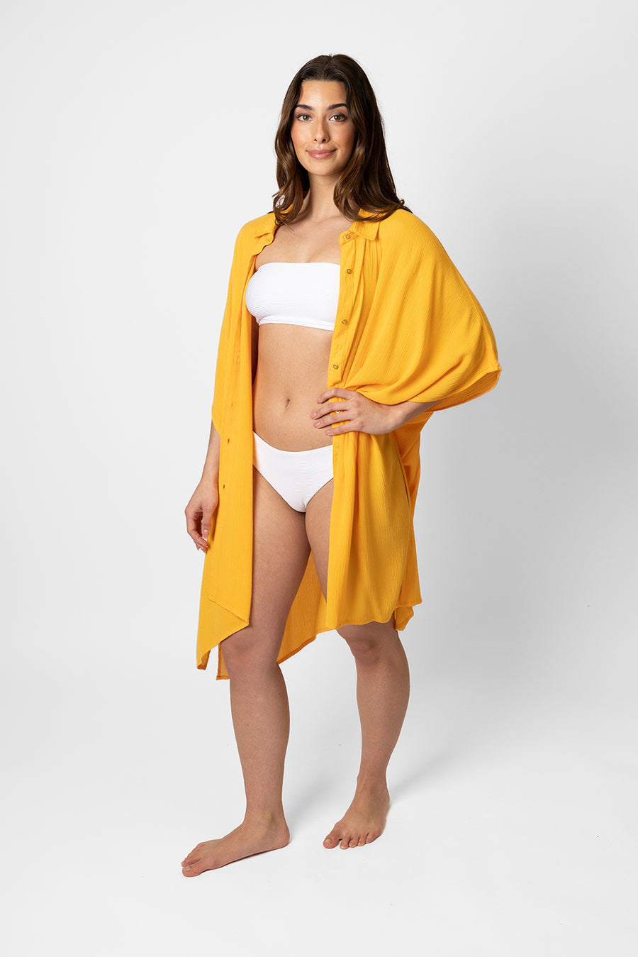 Brunette model facing side, wearing Yellow Mango Miami Big Shirt Button Up Dress. Features a structured collar, delicate button details, short sleeves, and flowy slits. Oversized and flowy with an asymmetrical hem, side slit, and back yoke. Perfect for breezy beach days. Koy Resort affordable vacation, cruise, and resort-wear.