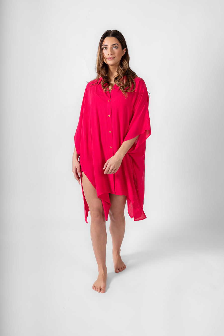 A front shot of a woman wearing a hot pink buttoned up shirt dress from Koy Resort. Brunette model facing front, wearing Raspberry Pink Miami Big Shirt. This oversized and flowy beach shirt dress features a structured collar, delicate button detail, and short sleeves. It has an asymmetrical hem, side slit, and back yoke, perfect for breezy beach days. Koy Resort affordable vacation, cruise, and resort-wear.