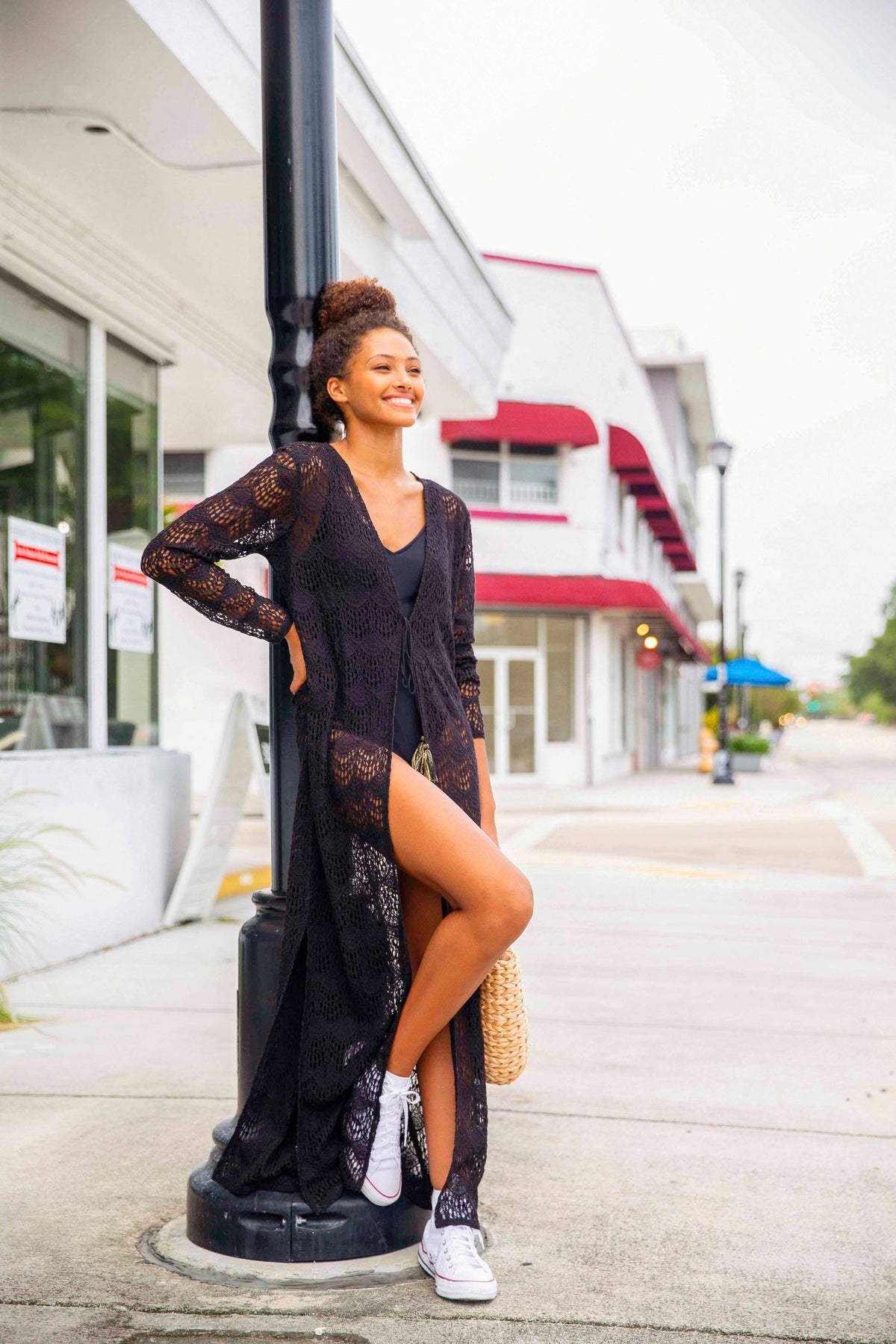 Model smiling wearing a long floor length black lace cardigan. Model posed leaning on a street lamp in Miami Florida.