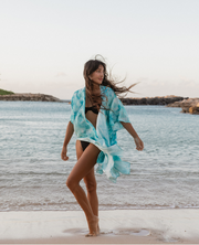 a women standing on the beach of Hawaii wearing a blue tie-dye kimono style cover up enjoying the ocean breeze