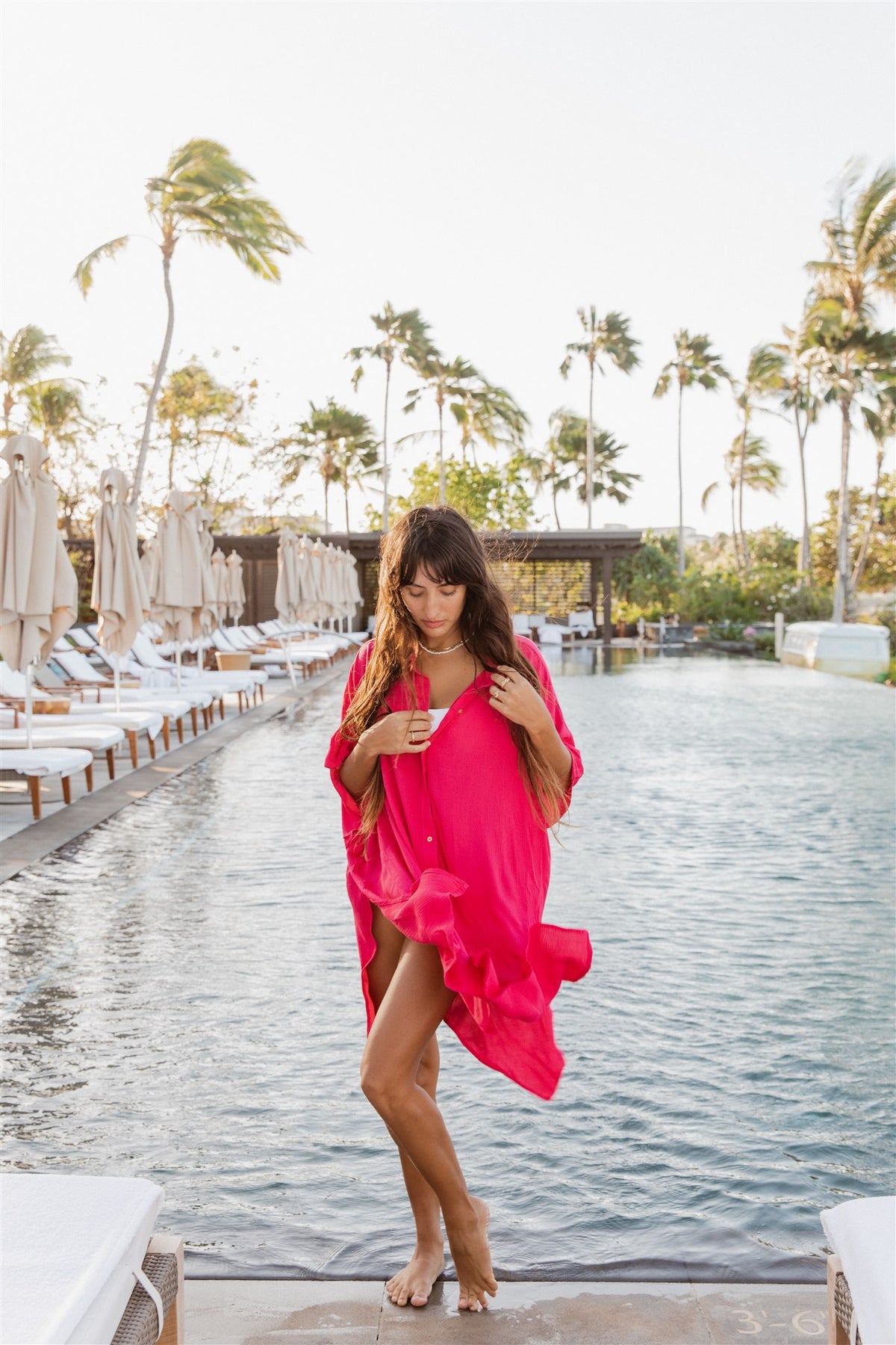 a woman wearing a hot pink shirt dress which flows with the ocean breeze standing in front of the waters