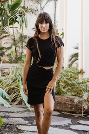 Brunette model facing front, wearing Black Laguna Rib Twisted Knot Top Mini Skirt with chic ribbed fabric, figure-flattering fit, knot top, side slit, and elastic back. Perfect for mix and match style and comfort. Koy Resort affordable vacation, cruise, and resort-wear.