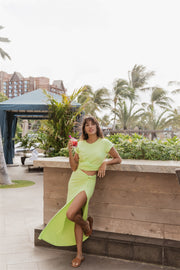 Brunette model facing far front, wearing the Margarita Green Laguna Rib Knot Top Maxi Skirt. This skirt features a ribbed finish, high dramatic slit, and a knot top design. Made from 92% Rayon and 8% Spandex, it's perfect for beach-to-street style. Koy Resort affordable vacation, cruise, and resort-wear.