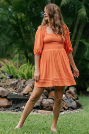 Blonde model facing front, wearing Orange Coral Punch Miami Smocked Mini Dress. Features a smocked bodice for a figure-flattering fit and puffy peasant-style sleeves for a bohemian touch. Perfect for the beach, a summer wedding, or a night out. Koy Resort affordable vacation, cruise, and resort-wear.sort-wear.