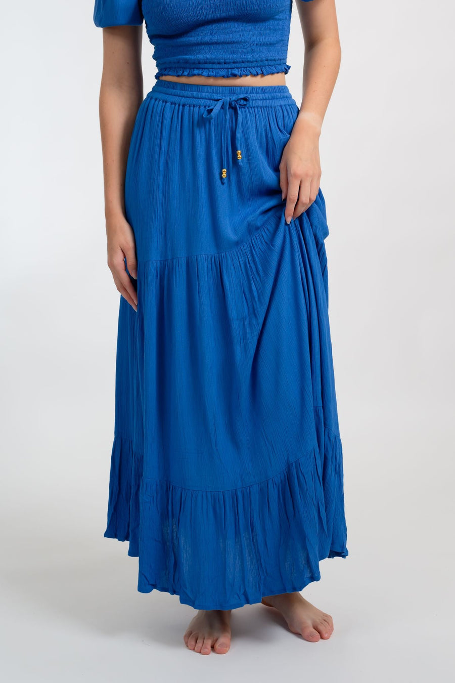 Blonde model facing front close up, wearing Blue Cobalt Miami Tiered Boho Maxi Skirt. Features a flowy tiered design, adjustable waistband with beaded ties, and side pockets. Crafted from crinkle rayon, it's perfect for bohemian beach days or romantic getaways. Koy Resort affordable vacation, cruise, and resort-wear.