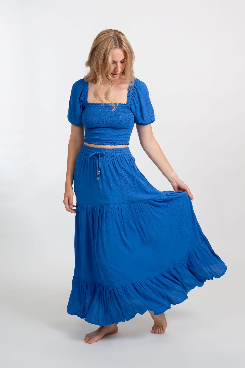 Blonde model facing front, wearing Blue Cobalt Miami Tiered Boho Maxi Skirt. Features a flowy tiered design, adjustable waistband with beaded ties, and side pockets. Crafted from crinkle rayon, it's perfect for bohemian beach days or romantic getaways. Koy Resort affordable vacation, cruise, and resort-wear.