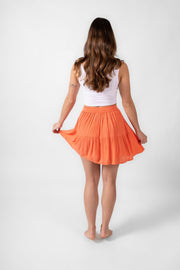 Brunette model facing back, wearing Coral Punch Orange Miami Tiered Mini Skirt. Features tiers with intricate crochet trim, an elastic waist for comfort and a flattering fit, and beaded detail with a metallic finish. Perfect for special occasions and adding playful flair to a fashion-forward vacation wardrobe. Koy Resort affordable vacation, cruise, and resort-wear.