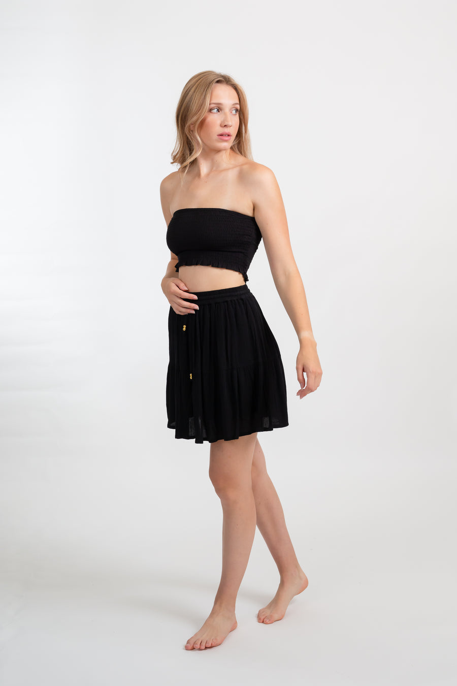 Blonde model facing side, wearing Black Miami Smocked Bandeau Top with a smocked elastic chest, cropped style, and bandeau design. Perfect for mixing and matching outfits. Koy Resort affordable vacation, cruise, and resort-wear.