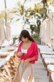 A brunette hair woman model wearing raspberry Miami collection tie front buttoned up top from Koy Resort and white Zuma Crochet Beach shorts from Koy Resort. Brunette model facing front, wearing a Raspberry Miami Tie Front Top. This vivacious top features chrome embellishments, a collar, and a tied knot in the front, blending sass and sophistication. It has a fitted top with a loose bodice, an adjustable front tie, and a button front. A best seller for daring yet sophisticated looks. Koy Resort affordable