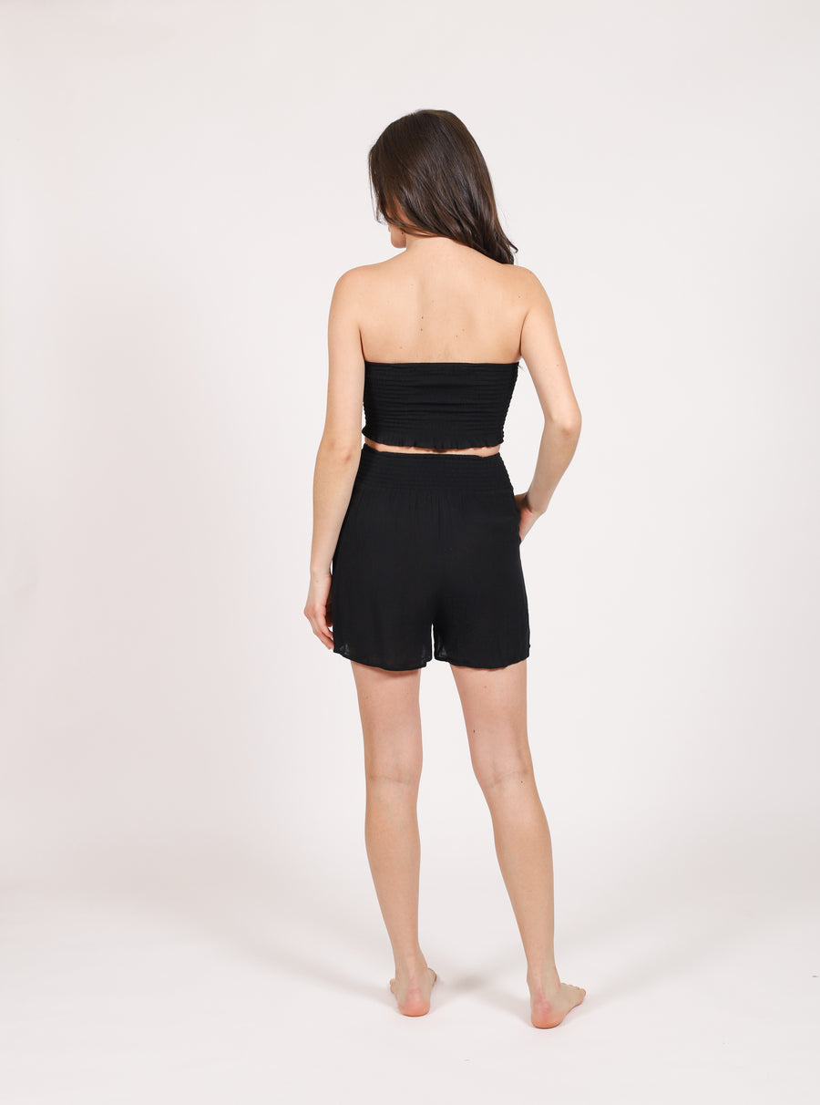 Brunette model facing back, wearing Black Miami Smocked Front Beach Shorts with a smocked band, side pockets, elastic waist, and flowy fit. Made from 100% rayon for a cool and comfortable feel. Koy Resort affordable vacation, cruise, and resort-wear.