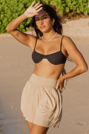 A confident woman in black bikini top and beige smocked beach shorts standing on the beach with on hand blocking the sun and the other hand on her waist. Brunette model facing front close up, wearing Coconut Miami Smocked Front Beach Shorts with a smocked band, side pockets, elastic waist, and flowy fit. Made from 100% rayon for a cool and comfortable feel. Koy Resort affordable vacation, cruise, and resort-wear.