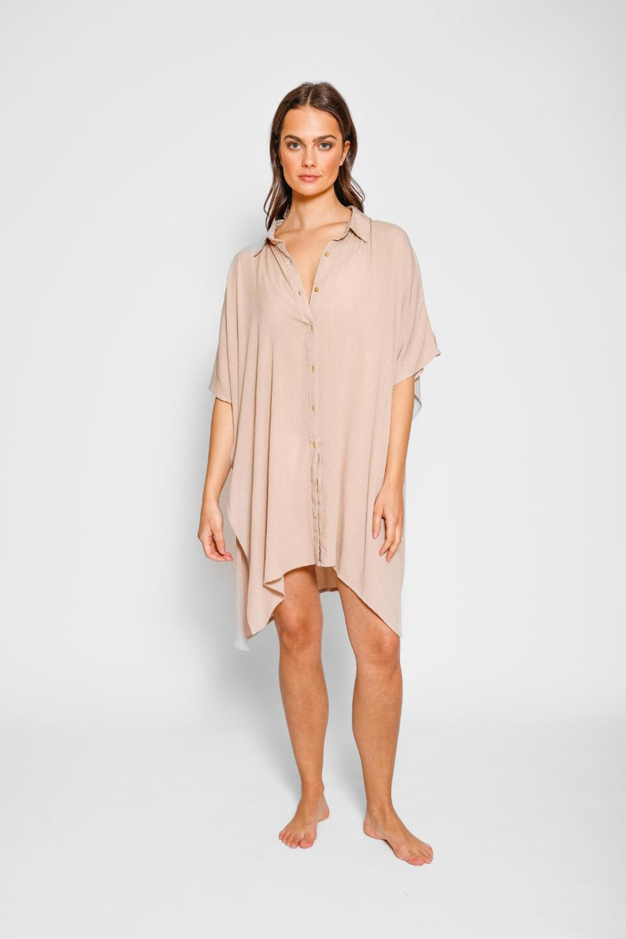 Brunette model facing front, wearing Coconut Miami Big Shirt Beach Dress with a structured collar, front button-up, short sleeves, asymmetrical hem, side slit, and back yoke. Oversized and flowy for breezy beach days. Koy Resort affordable vacation, cruise, and resort-wear.