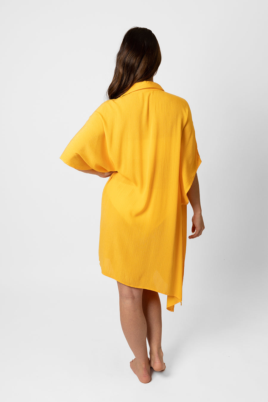 Brunette model facing back, wearing Yellow Mango Miami Big Shirt Button Up Dress. Features a structured collar, delicate button details, short sleeves, and flowy slits. Oversized and flowy with an asymmetrical hem, side slit, and back yoke. Perfect for breezy beach days. Koy Resort affordable vacation, cruise, and resort-wear.