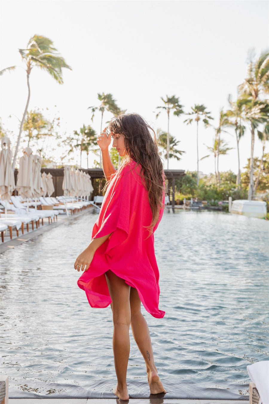 Brunette model facing back, wearing Raspberry Pink Miami Big Shirt. This oversized and flowy beach shirt dress features a structured collar, delicate button detail, and short sleeves. It has an asymmetrical hem, side slit, and back yoke, perfect for breezy beach days. Koy Resort affordable vacation, cruise, and resort-wear.