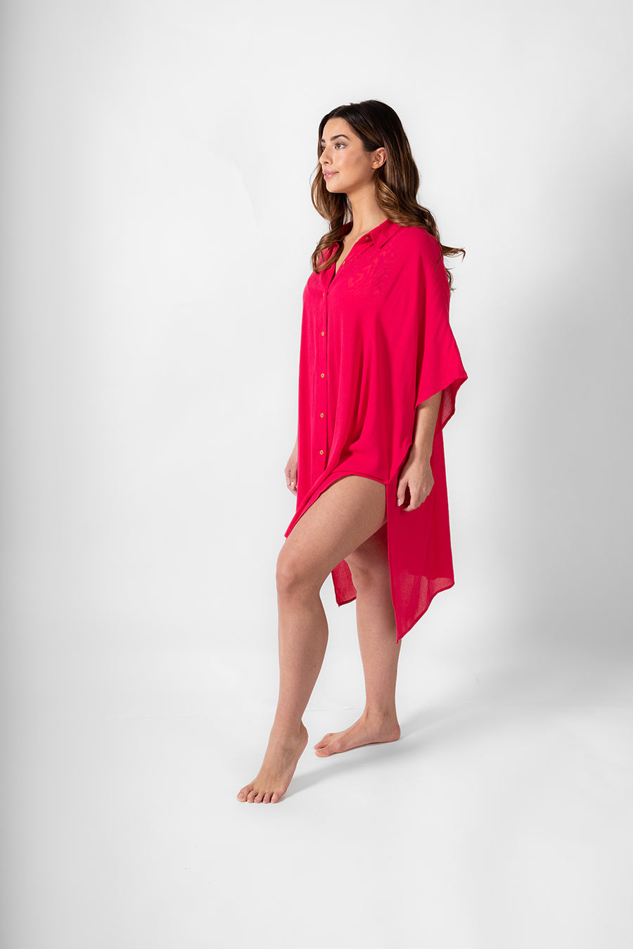 A side shot of a woman wearing a hot pink shirt dress from Koy Resort being confident. Brunette model facing side, wearing Raspberry Pink Miami Big Shirt. This oversized and flowy beach shirt dress features a structured collar, delicate button detail, and short sleeves. It has an asymmetrical hem, side slit, and back yoke, perfect for breezy beach days. Koy Resort affordable vacation, cruise, and resort-wear.