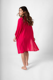 A back of a woman wearing a hot pink shirt dress. Brunette model facing back, wearing Raspberry Pink Miami Big Shirt. This oversized and flowy beach shirt dress features a structured collar, delicate button detail, and short sleeves. It has an asymmetrical hem, side slit, and back yoke, perfect for breezy beach days. Koy Resort affordable vacation, cruise, and resort-wear.