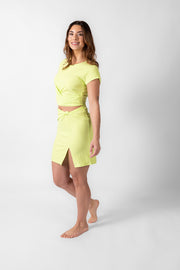 Brunette model facing side, wearing the Margarita Green Laguna Rib Twisted Crop Top. The top features short sleeves, rib fabric, a twist hem, and a crew neck, offering a chic and flattering fit. Made from 92% Rayon and 8% Spandex. Available in black or striking lime margarita. Koy Resort affordable vacation, cruise, and resort-wear.