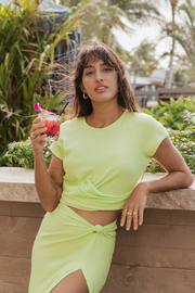 Laguna Rib Twisted Crop Top - Lime margarita. Model holding tropical cocktail, wearing laguna rib crop top on vacation at tropical destination resort hotel in Hawaii. Brunette model facing front, wearing the Margarita Green Laguna Rib Twisted Crop Top. The top features short sleeves, rib fabric, a twist hem, and a crew neck, offering a chic and flattering fit. Made from 92% Rayon and 8% Spandex. Available in black or striking lime margarita. Koy Resort affordable vacation, cruise, and resort-wear.