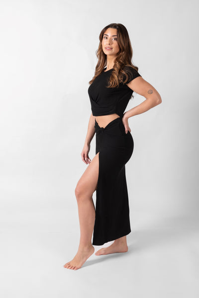 Brunette model facing side, wearing Black Laguna Rib Knot Top Maxi Skirt with ribbed cotton fabric, high dramatic slit, and knot top. Perfect for beach-to-street style and mix-and-match outfits. Koy Resort affordable vacation, cruise, and resort-wear.