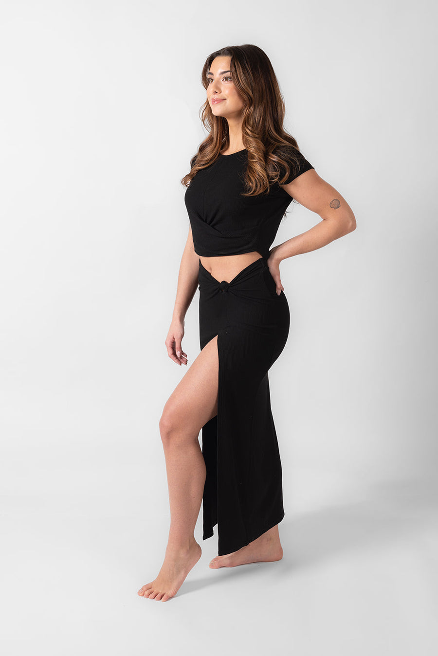 Laguna Rib Twisted Crop Top in black color. Brunette model facing side, wearing Black Laguna Rib Twisted Crop Top with short sleeves, ribbed fabric, twist hem, and crew neck. Flattering and chic for beach-to-street transitions. Perfect for pairing with the Laguna Rib Mini Skirt or Knot Maxi Skirt. Koy Resort affordable vacation, cruise, and resort-wear.