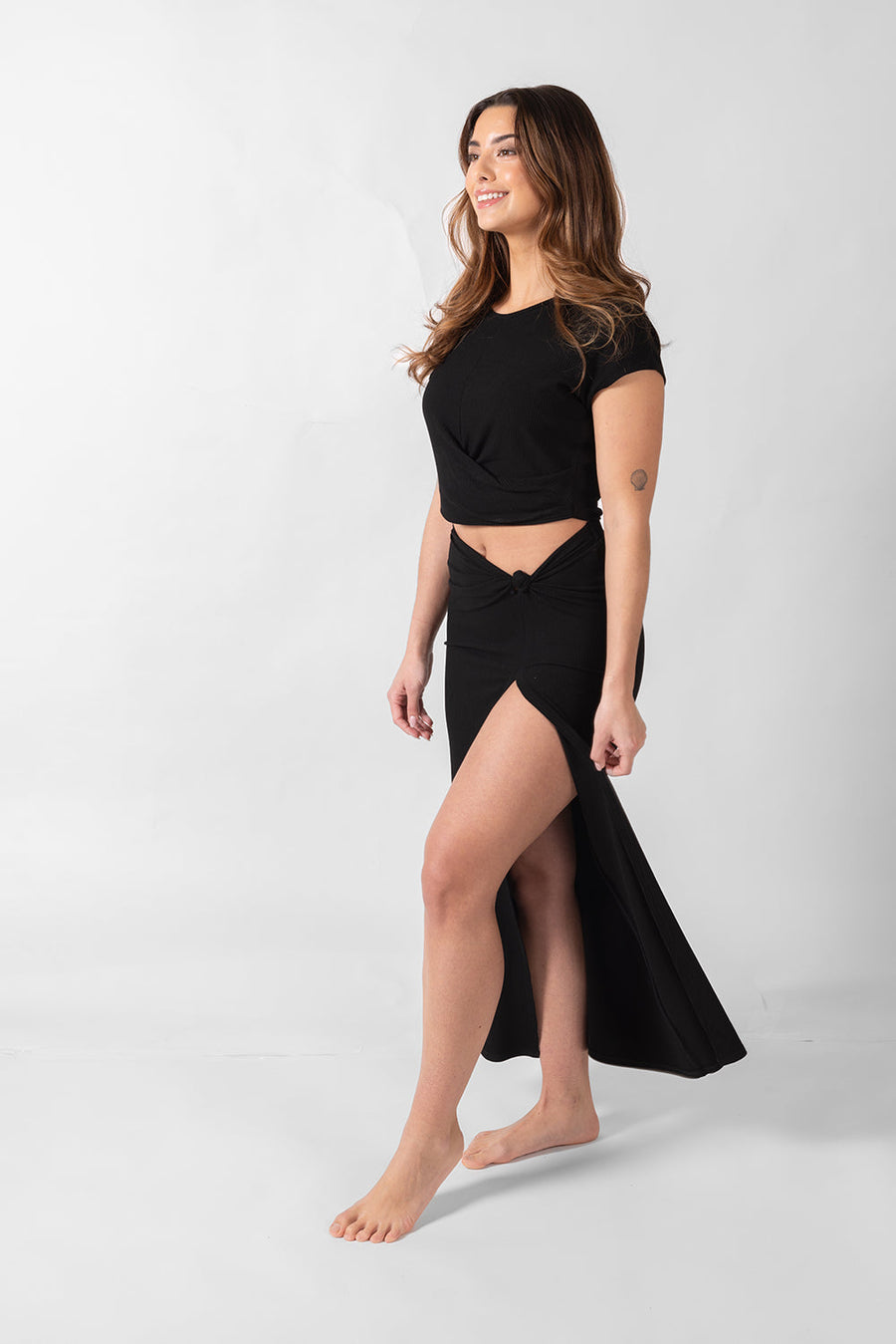 Brunette model facing side, wearing Black Laguna Rib Knot Top Maxi Skirt with ribbed cotton fabric, high dramatic slit, and knot top. Perfect for beach-to-street style and mix-and-match outfits. Koy Resort affordable vacation, cruise, and resort-wear.