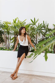 Brunette model facing side, wearing Black Laguna Rib Twisted Knot Top Mini Skirt with chic ribbed fabric, figure-flattering fit, knot top, side slit, and elastic back. Perfect for mix and match style and comfort. Koy Resort affordable vacation, cruise, and resort-wear.