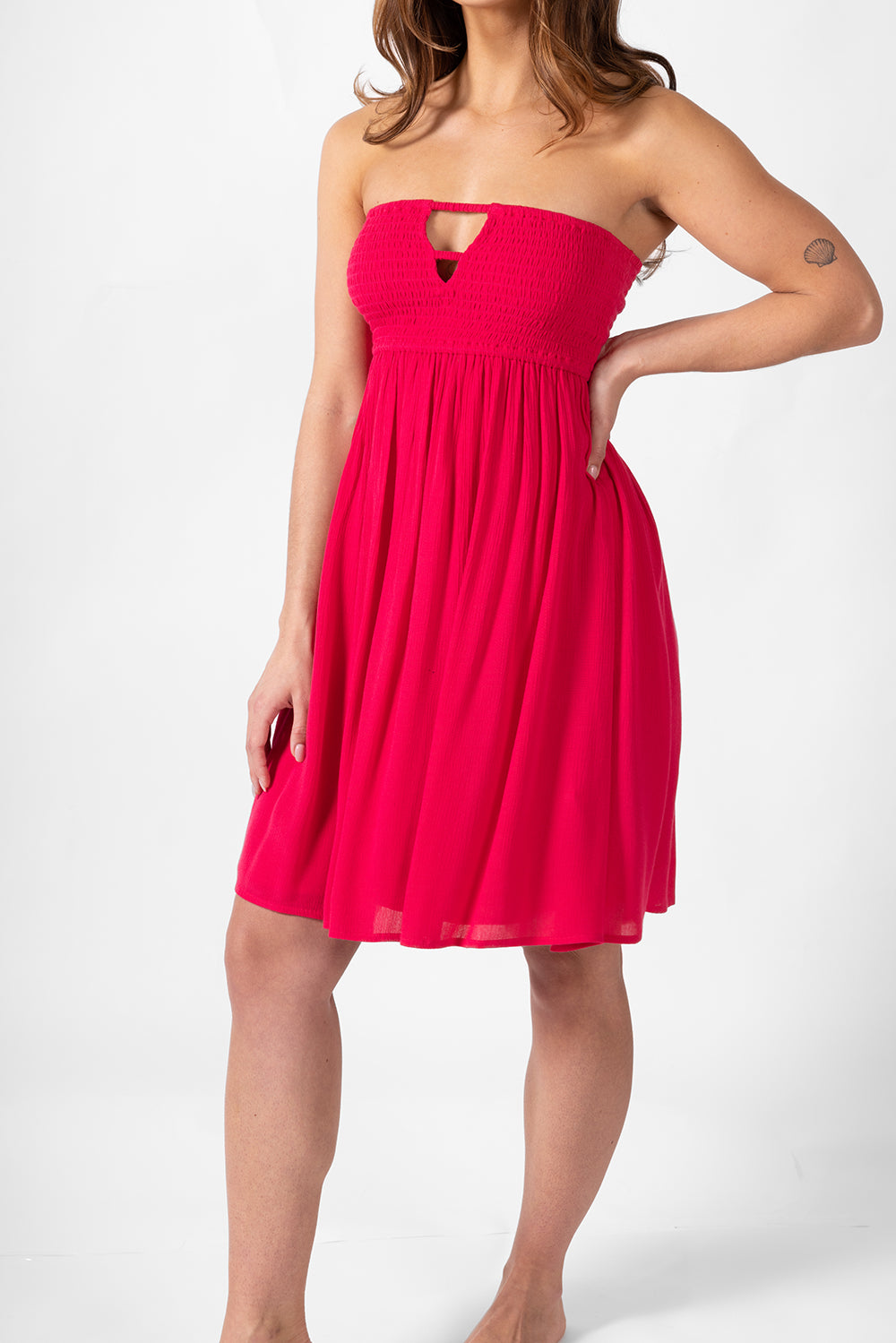 A woman model wearing a hot pink smocked bandeau mini dress with one hand on her waist. Brunette model facing side close up, wearing Raspberry Pink Miami Strapless Bandeau Mini Dress. This loose and flowy dress features an elastic bandeau top with a peek-a-boo bodice, and side slits for a flirty look. The dress is lined in white and has an adjustable cord at the back. Koy Resort affordable vacation, cruise, and resort-wear.