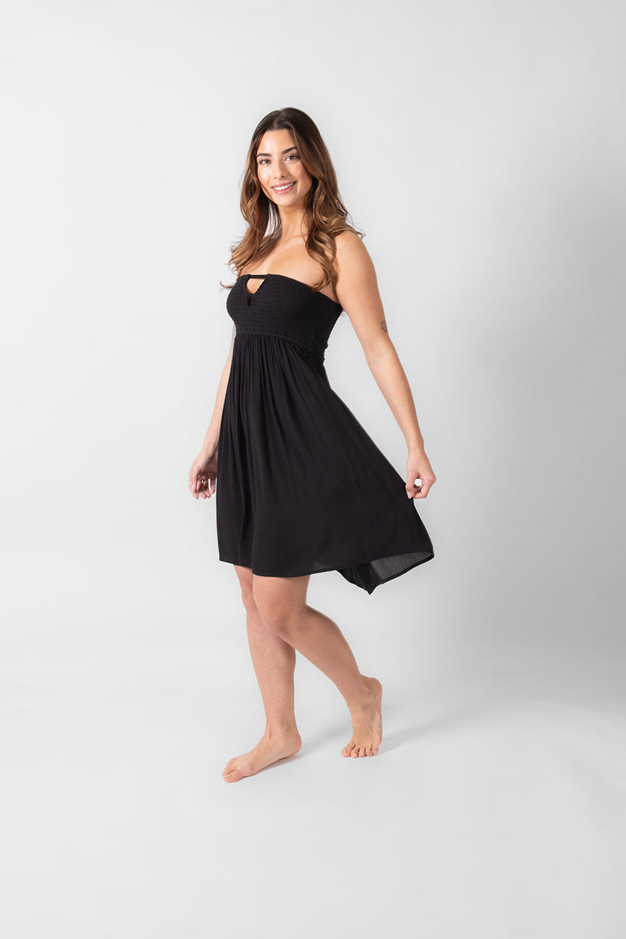 A side shot of a dark hair woman in a black smocked bandeau mini dress holding smiling. Brunette model facing front, wearing Black Miami Strapless Bandeau Mini Dress with an elastic bandeau, peek-a-boo bodice, and mini length. Made from rayon fabric for an effortless yet flirty look. Koy Resort affordable vacation, cruise, and resort-wear.