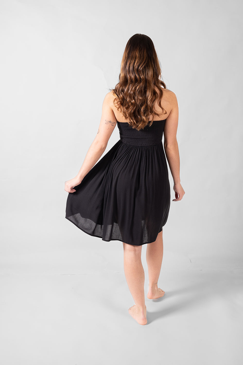 the back of a dark hair woman in a black bandeau beach mini dress with one hand holding the edge of the dress 