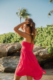 Brunette model facing side close up, wearing Raspberry Pink Miami Strapless Bandeau Mini Dress. This loose and flowy dress features an elastic bandeau top with a peek-a-boo bodice, and side slits for a flirty look. The dress is lined in white and has an adjustable cord at the back. Koy Resort affordable vacation, cruise, and resort-wear.