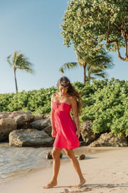 A woman in a hot pink smocked bandeau mini dress standing on the sand of the beach in Hawaii with palm trees and ocean breeze in the background. Brunette model facing side, wearing Raspberry Pink Miami Strapless Bandeau Mini Dress. This loose and flowy dress features an elastic bandeau top with a peek-a-boo bodice, and side slits for a flirty look. The dress is lined in white and has an adjustable cord at the back. Koy Resort affordable vacation, cruise, and resort-wear.