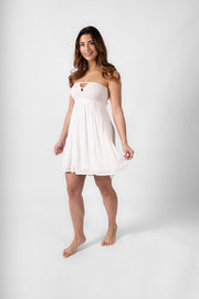 A woman in a white smocked bandeau white mini beach dress. Brunette model facing front, wearing White Miami Strapless Bandeau Mini Dress with an elastic bandeau, peek-a-boo bodice, and mini length. Made from rayon fabric for an effortless yet flirty look. Koy Resort affordable vacation, cruise, and resort-wear.