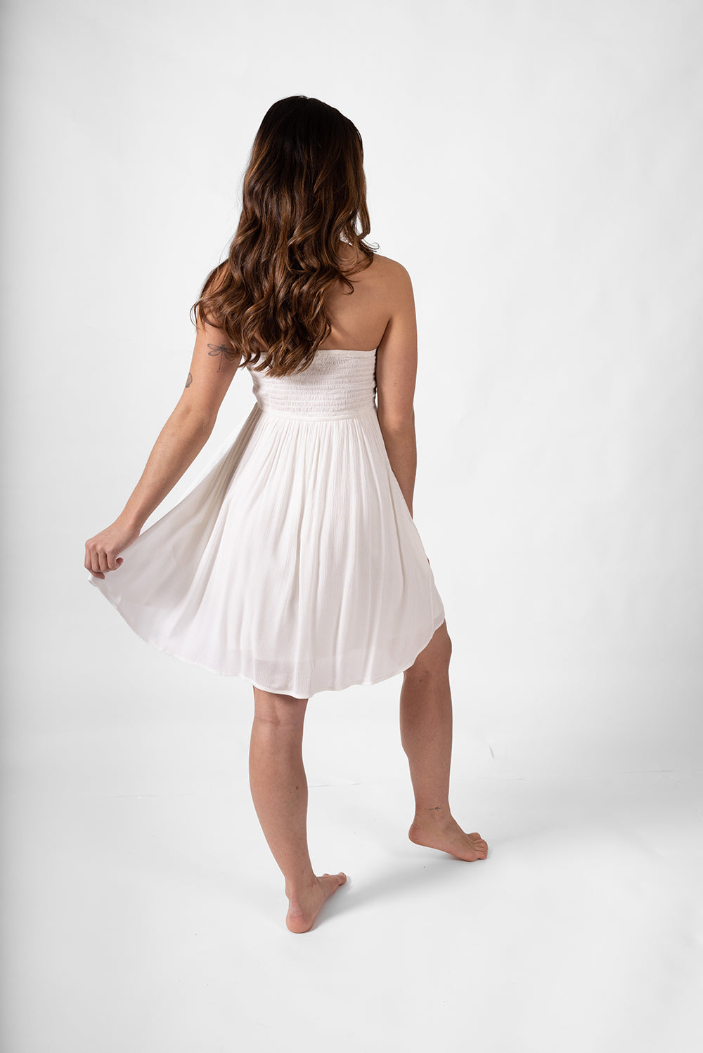 the back of a brunette woman model wearing a white beachy bandeau mini dress with one hand on the edge of the dress to show how flowy it is