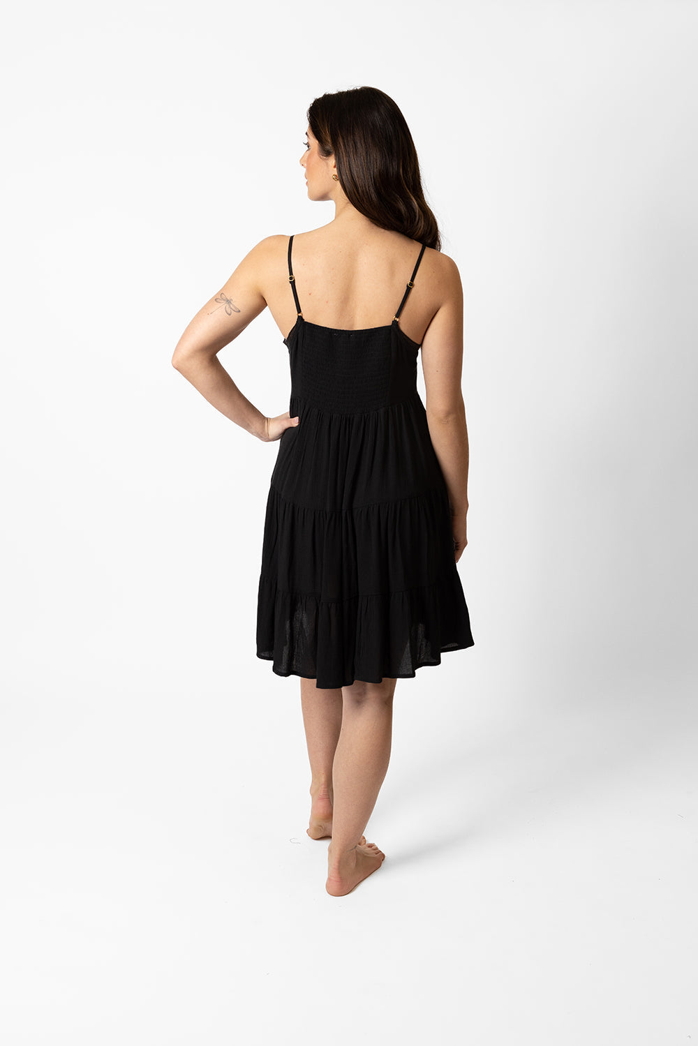 the back of a dark hair woman wearing a black spaghetti strap beach mini dress with one hand on her waist