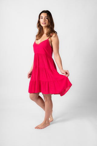 a brunette hair woman standing on the side angle holding the edge of her raspberry color tiered fabric strappy mini dress