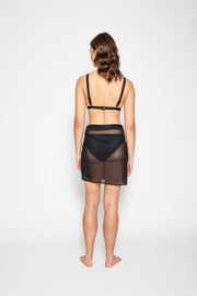 Brunette model facing back, wearing the Black Escape Mesh Mini Sarong Skirt. This versatile mini sarong is crafted from exclusive mesh fabric, ideal for beach or pool days. The semi-sheer design offers a trendy, effortless look, with multiple tying options for a personalized style. Koy Resort affordable vacation, cruise, and resort-wear.