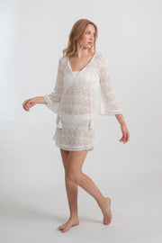 Blonde model facing front, wearing the Cream Flamenco Lace Crochet Beach Cover Up Tunic. This elegant cover-up features delicate crochet detailing, bell sleeves with lace trim, fringe hem, and long sleeves with tassel detail. Made from 55% Cotton and 45% Nylon. Koy Resort affordable vacation, cruise, and resort-wear.