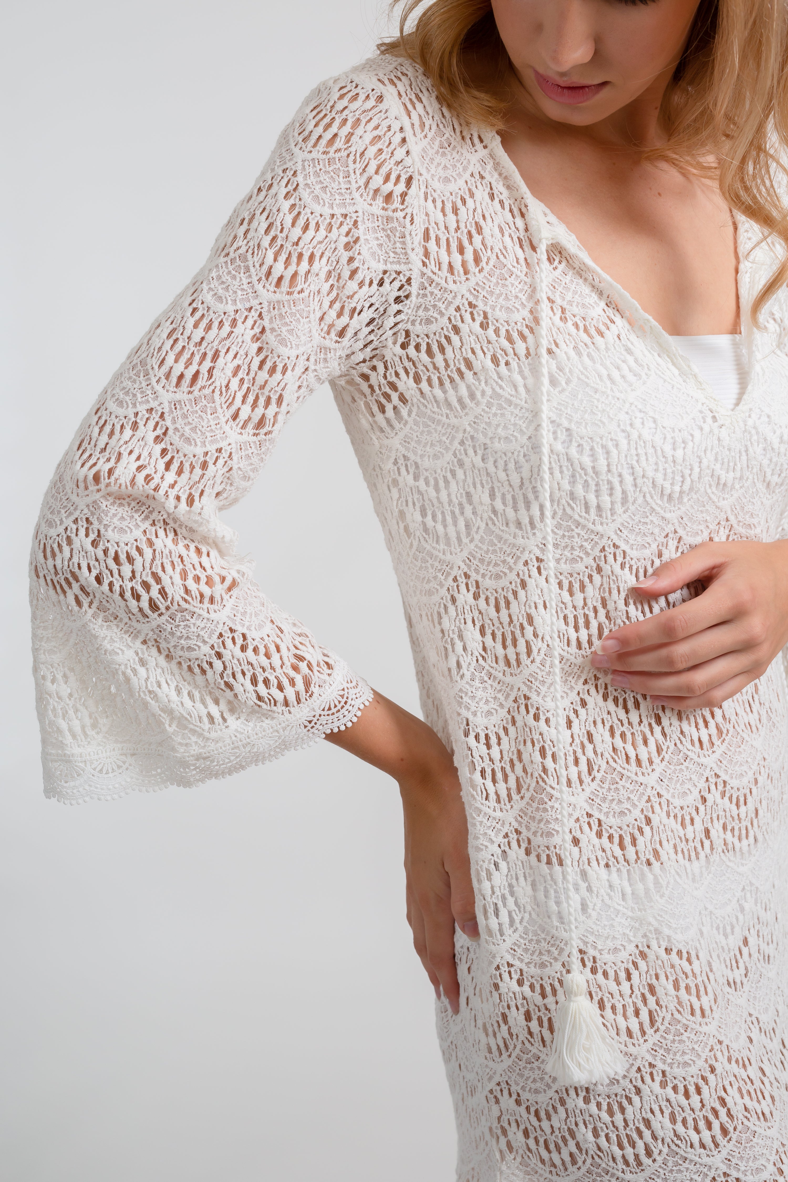 Blonde model facing front close up, wearing the Cream Flamenco Lace Crochet Beach Cover Up Tunic. This elegant cover-up features delicate crochet detailing, bell sleeves with lace trim, fringe hem, and long sleeves with tassel detail. Made from 55% Cotton and 45% Nylon. Koy Resort affordable vacation, cruise, and resort-wear.