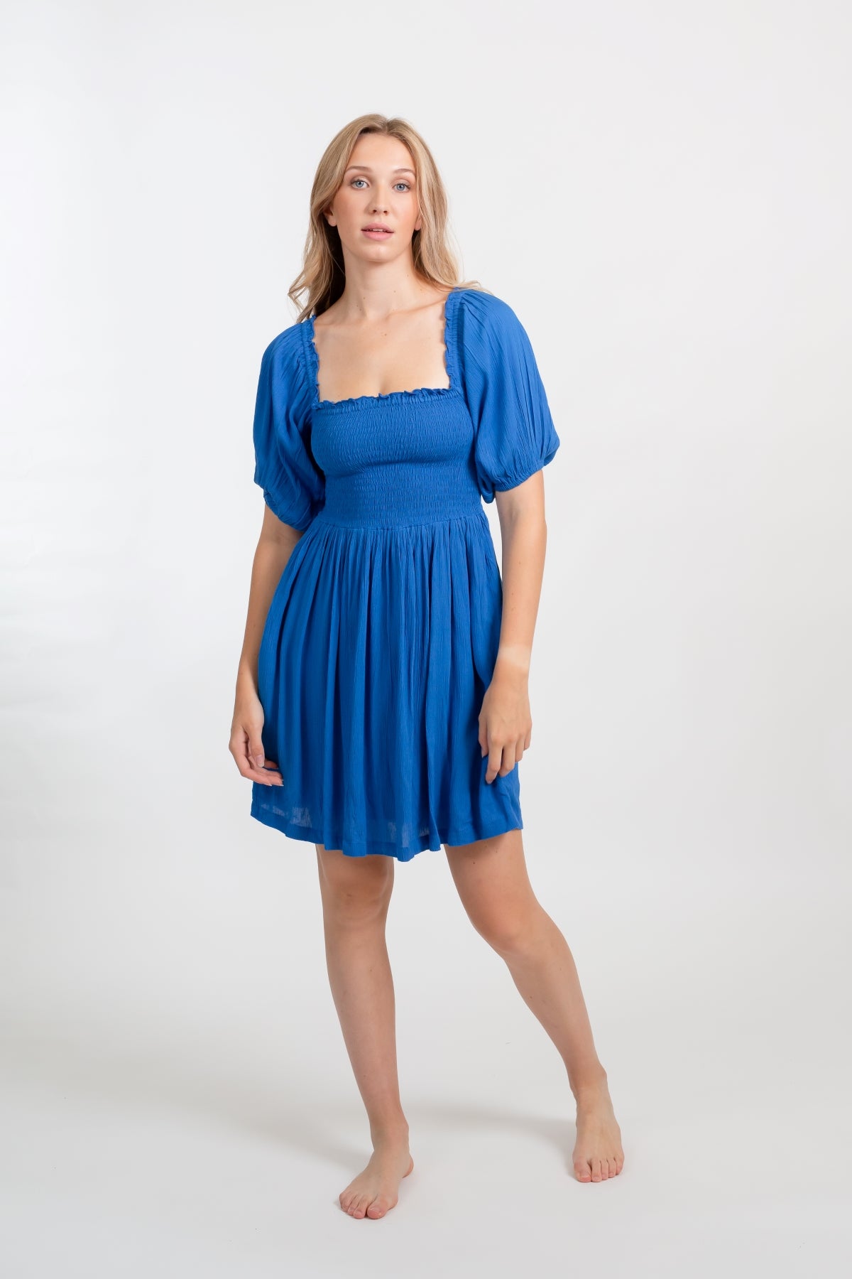 Blonde model facing front, wearing Blue Cobalt Miami Smocked Mini Dress. Features a smocked bodice for a figure-flattering fit and puffy peasant-style sleeves for a bohemian touch. Perfect for the beach, a summer wedding, or a night out. Koy Resort affordable vacation, cruise, and resort-wear.sort-wear.