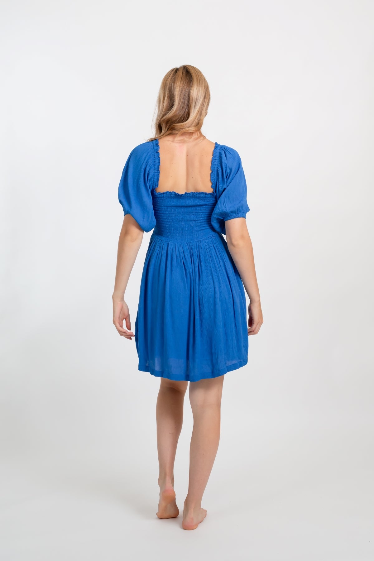 Blonde model facing back, wearing Blue Cobalt Miami Smocked Mini Dress. Features a smocked bodice for a figure-flattering fit and puffy peasant-style sleeves for a bohemian touch. Perfect for the beach, a summer wedding, or a night out. Koy Resort affordable vacation, cruise, and resort-wear.sort-wear.