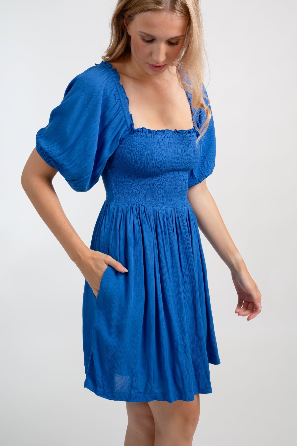 Blonde model facing side close up, wearing Blue Cobalt Miami Smocked Mini Dress. Features a smocked bodice for a figure-flattering fit and puffy peasant-style sleeves for a bohemian touch. Perfect for the beach, a summer wedding, or a night out. Koy Resort affordable vacation, cruise, and resort-wear.sort-wear.