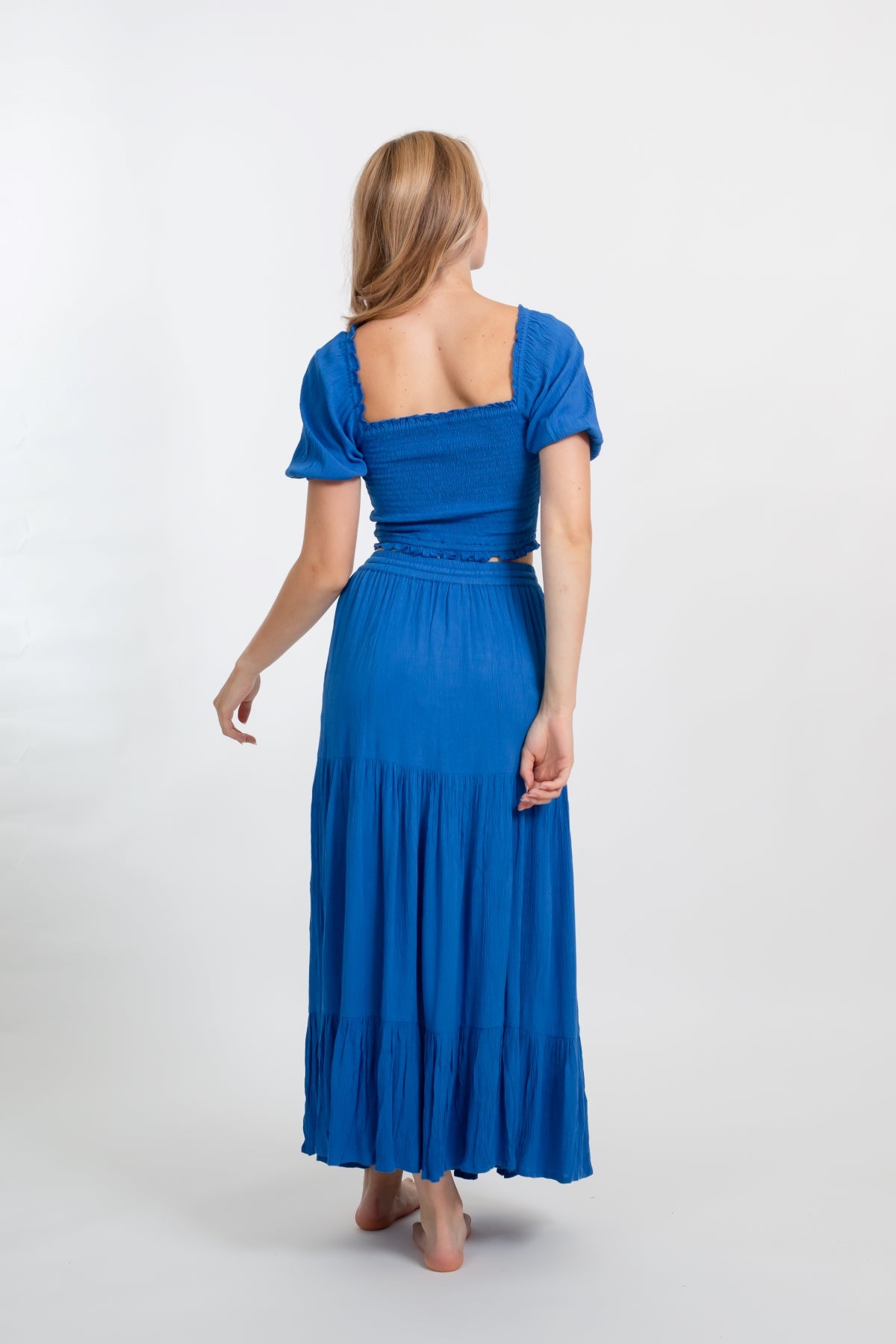 Blonde model facing back, wearing Blue Cobalt Miami Tiered Boho Maxi Skirt. Features a flowy tiered design, adjustable waistband with beaded ties, and side pockets. Crafted from crinkle rayon, it's perfect for bohemian beach days or romantic getaways. Koy Resort affordable vacation, cruise, and resort-wear.