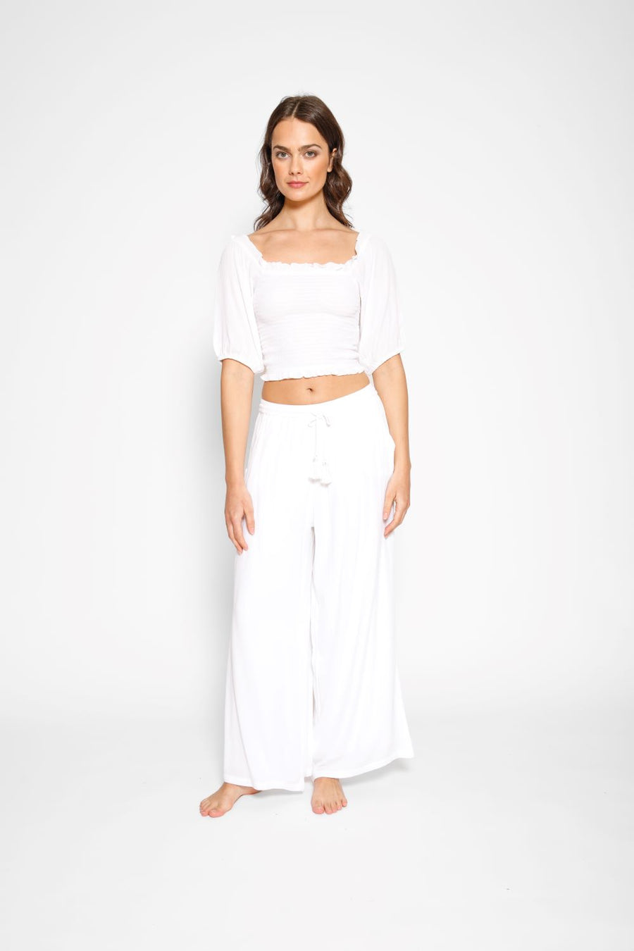 Brunette model facing front, wearing White Miami Wide Leg Pant with a tie waist, palazzo style, flowy straight leg, elastic waist, side pockets, and metallic tassel drawcord. Koy Resort affordable vacation, cruise, and resort-wear.