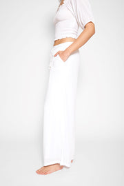 Brunette model facing side, wearing White Miami Wide Leg Pant with a tie waist, palazzo style, flowy straight leg, elastic waist, side pockets, and metallic tassel drawcord. Koy Resort affordable vacation, cruise, and resort-wear.