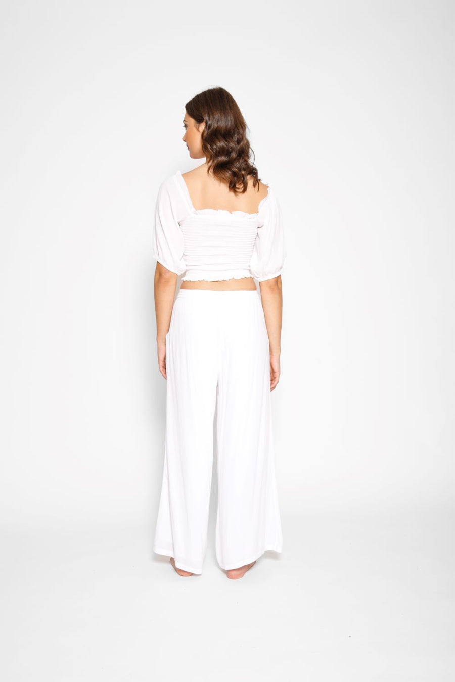 Brunette model facing back, wearing White Miami Wide Leg Pant with a tie waist, palazzo style, flowy straight leg, elastic waist, side pockets, and metallic tassel drawcord. Koy Resort affordable vacation, cruise, and resort-wear.