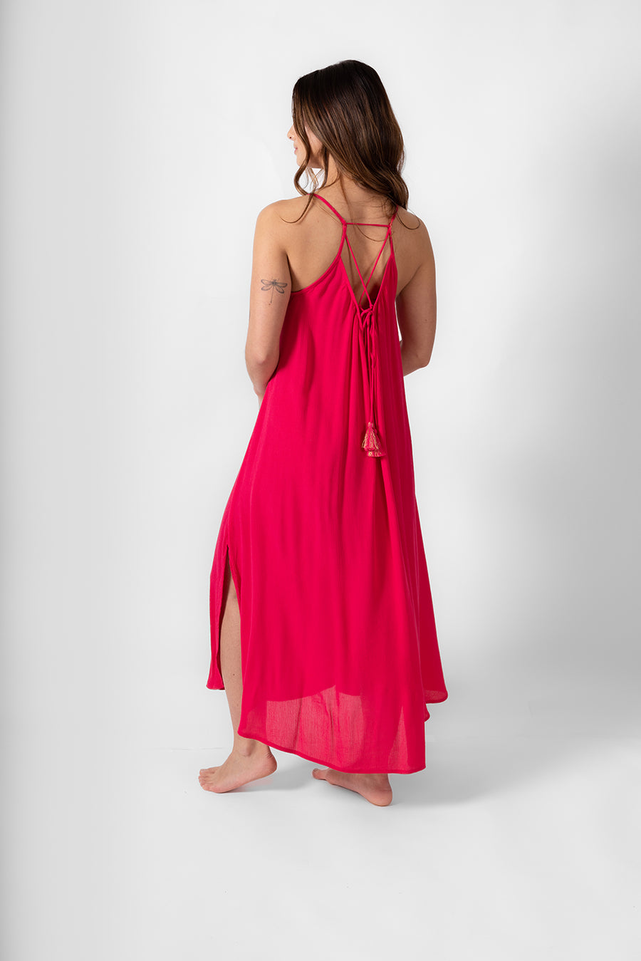 The back side of the a brunette model wearing raspberry colored strappy flowy midi dress with a side slip up to the knee. Brunette model facing front, wearing Raspberry Pink Miami Side Slit Strappy Midi Dress. The dress features a strappy silhouette, midi length, and breezy material, with side slits and an adjustable cord at the back. The dress is lined in white, offering a loose and flowy fit perfect for effortless, fashion-forward looks. Koy Resort affordable vacation, cruise, and resort-wear.