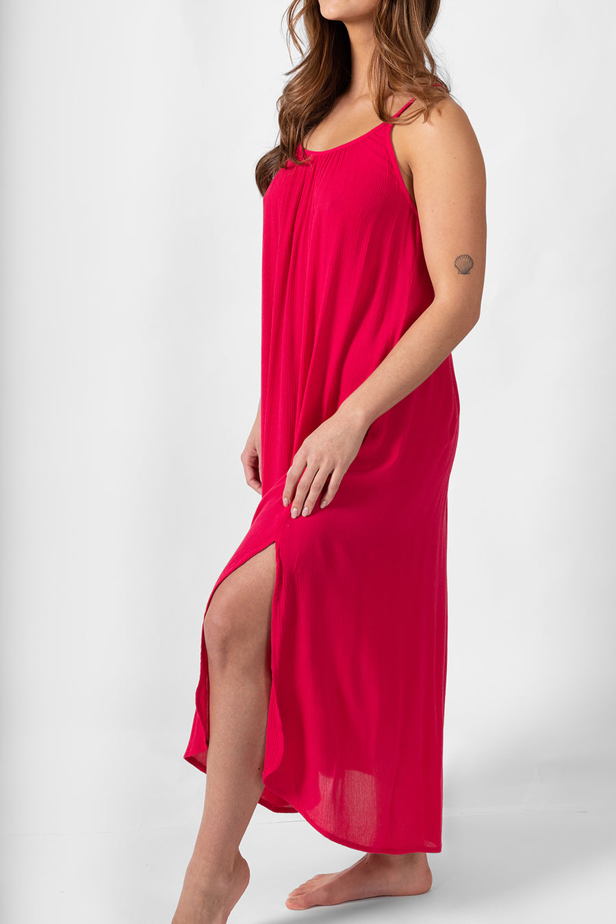 A side shot of a woman model wearing a side slipt strappy midi dress in raspberry color. Brunette model facing front, wearing Raspberry Pink Miami Side Slit Strappy Midi Dress. The dress features a strappy silhouette, midi length, and breezy material, with side slits and an adjustable cord at the back. The dress is lined in white, offering a loose and flowy fit perfect for effortless, fashion-forward looks. Koy Resort affordable vacation, cruise, and resort-wear.