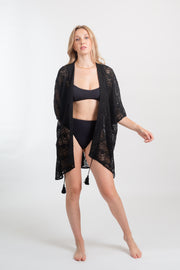 Blonde model facing front, wearing the Black Flamenco Lace Crochet Beach Cover Up Kimono. This stylish cover-up features an open front, scoop hem, long tie-up tassel, short sleeves, and sheer crochet lace detailing. Made from 55% Cotton and 45% Nylon. Koy Resort affordable vacation, cruise, and resort-wear.