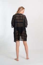 Blonde model facing back, wearing the Black Flamenco Lace Crochet Beach Cover Up Kimono. This stylish cover-up features an open front, scoop hem, long tie-up tassel, short sleeves, and sheer crochet lace detailing. Made from 55% Cotton and 45% Nylon. Koy Resort affordable vacation, cruise, and resort-wear.